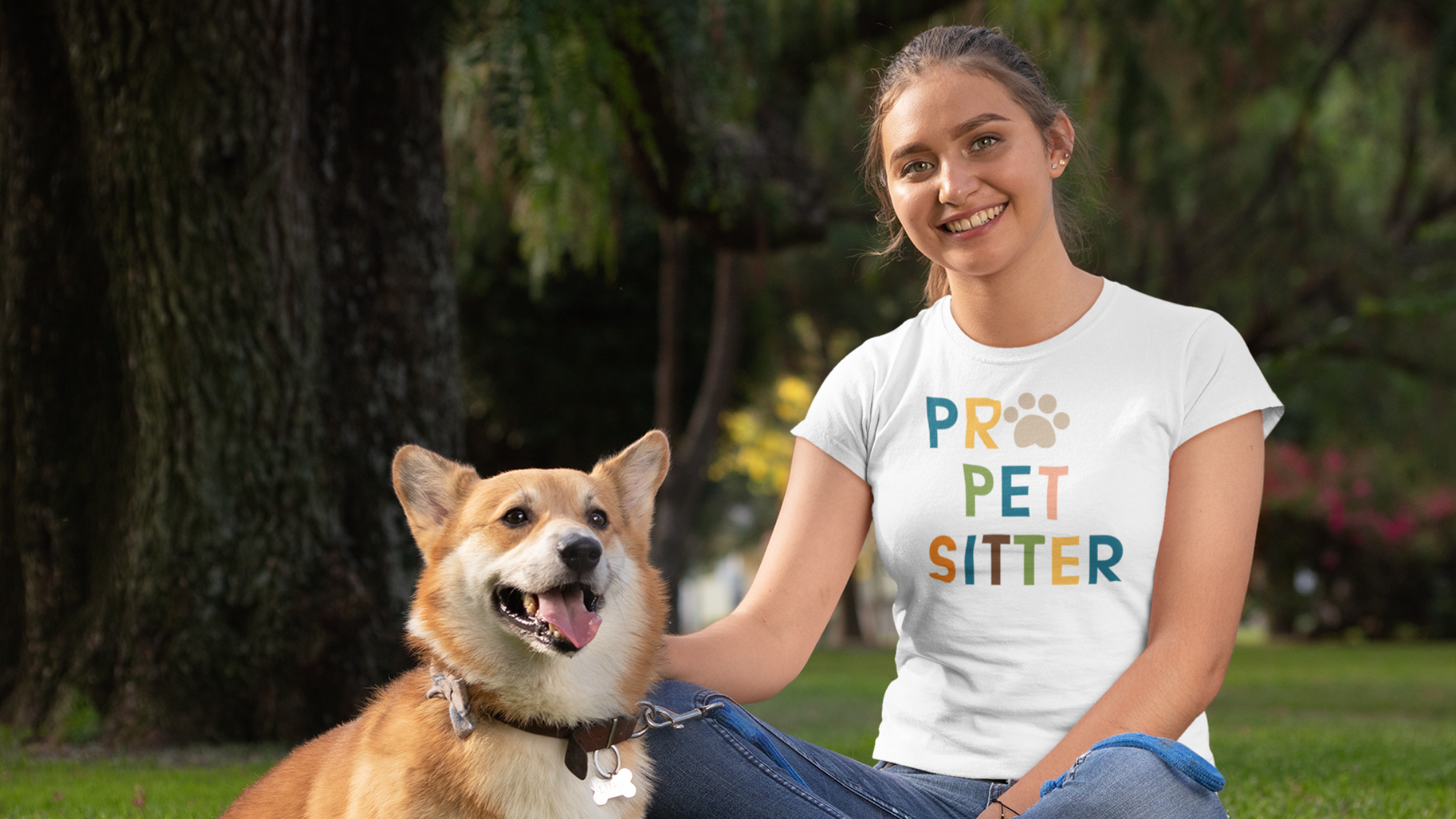 Young adult woman wearing denim jeans and a white t-shirt that says PRO PET SITTER sitting in the grass with a Corgi dog with brown and white fur